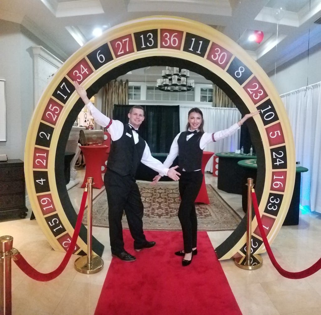 Card dealers standing on red carpet by roulette wheel entry to casino themed party